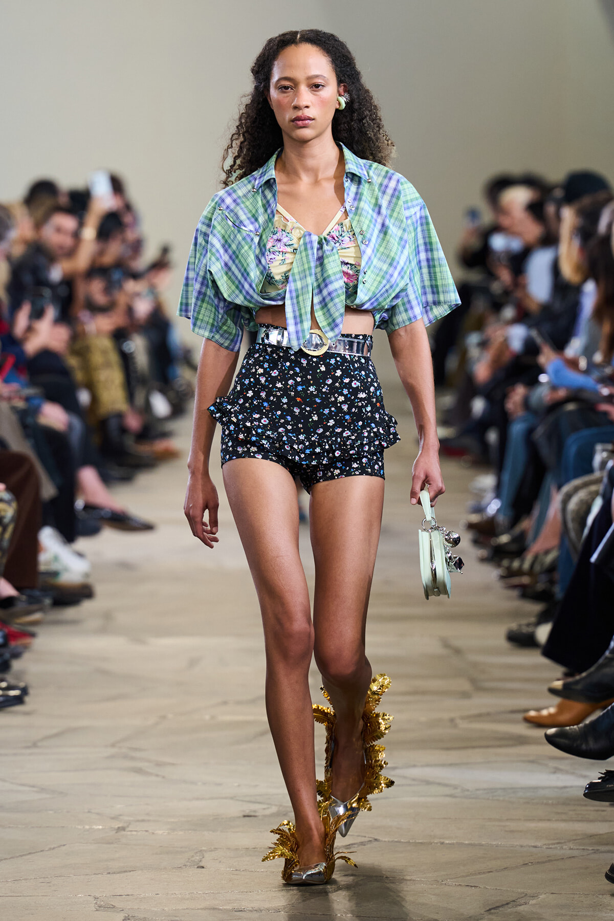 Model walking a runway in a green plaid tied blouse over a floral top, black floral shorts, and a metallic belt with gold-leaf heels on both feet.