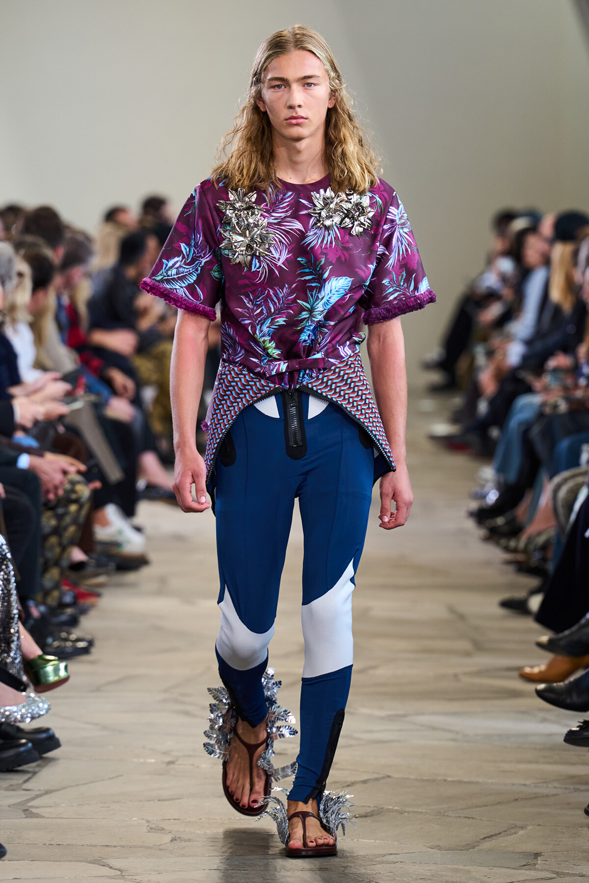 Model walks a fashion show wearing a purple tropical-print top with metallic appliqués and blue color-block pants with silver fringe sandals.