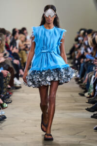 Model walks a fashion runway wearing a sky-blue sleeveless dress with fringe at the shoulders and a metallic ruffled silver hem, plus oversized protective goggles and sandals.