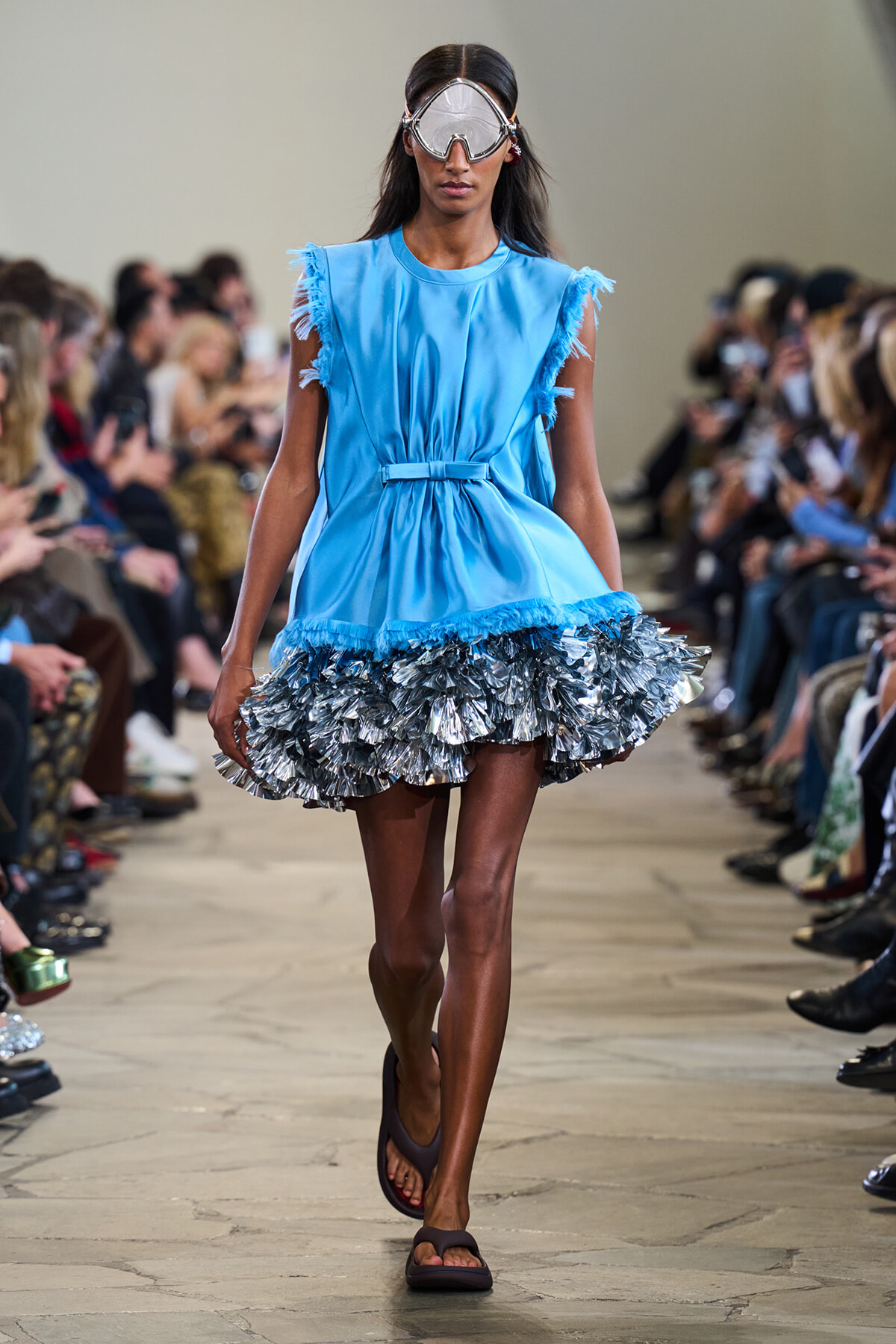 Model walks a fashion runway wearing a sky-blue sleeveless dress with fringe at the shoulders and a metallic ruffled silver hem, plus oversized protective goggles and sandals.