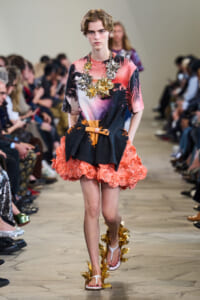 Model walks a runway in a tropical-print dress with a coral fringe skirt, large gold leaf necklace, and matching sandals.