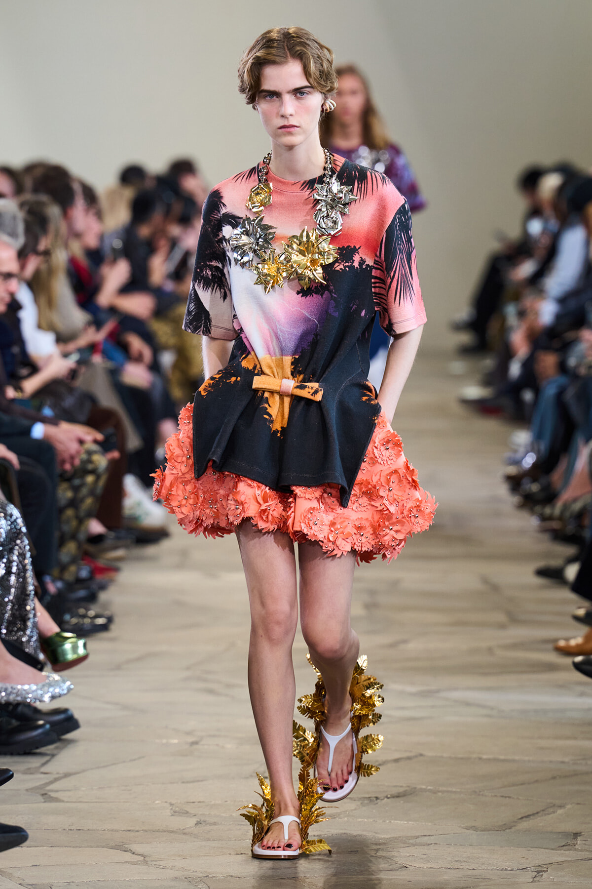 Model walks a runway in a tropical-print dress with a coral fringe skirt, large gold leaf necklace, and matching sandals.