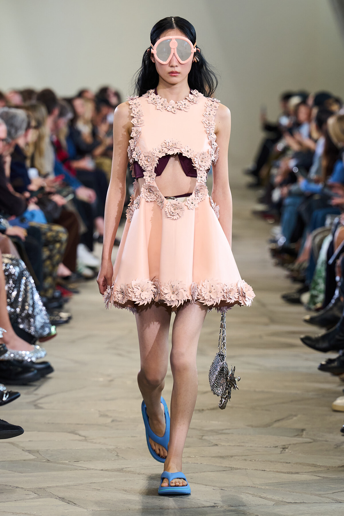 Model on a runway wearing a pale pink, flower-embellished mini dress with cutouts, oversized pink goggles, blue sandals, and a metallic bag.