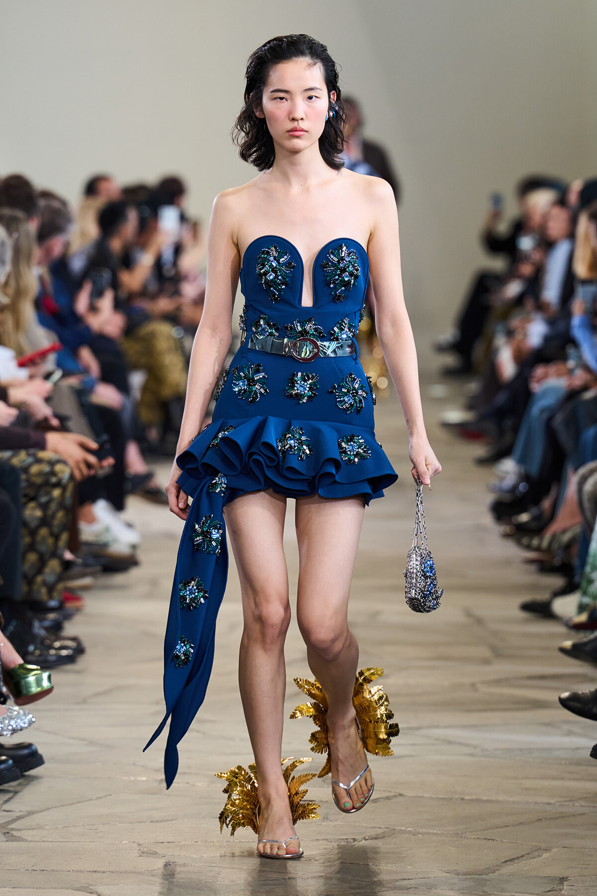 Model walks a blue strapless dress with embroidered embellishments along the bodice and hem, on a runway lined with spectators.
