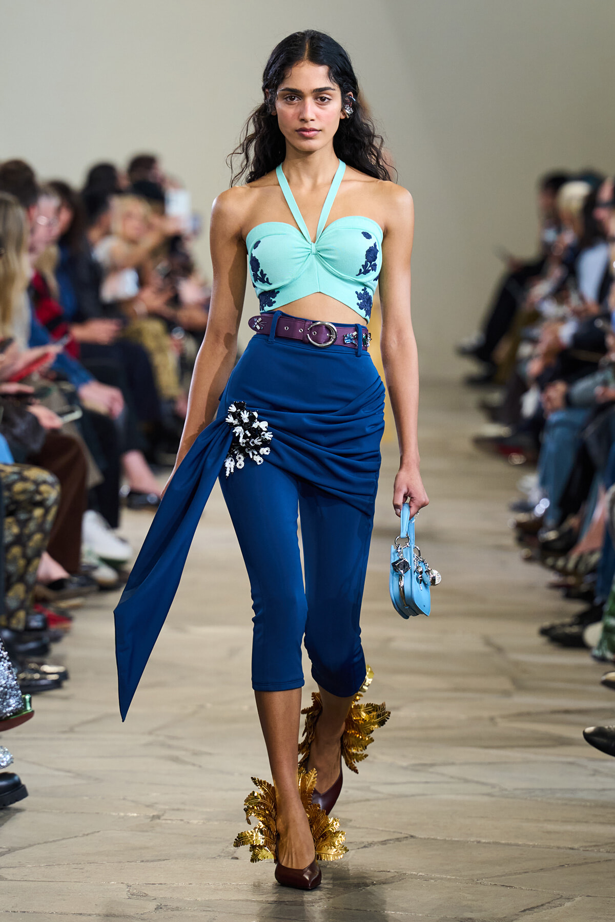 Model walking a runway in a turquoise halter crop top and high-waisted blue pants with a draped overlay and floral embellishment, carrying a small blue handbag with charms.