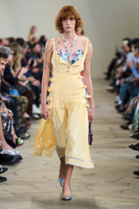 Model walking a runway in a pale yellow dress with a floral bustier top and side buckle details, audience blurred in the background.