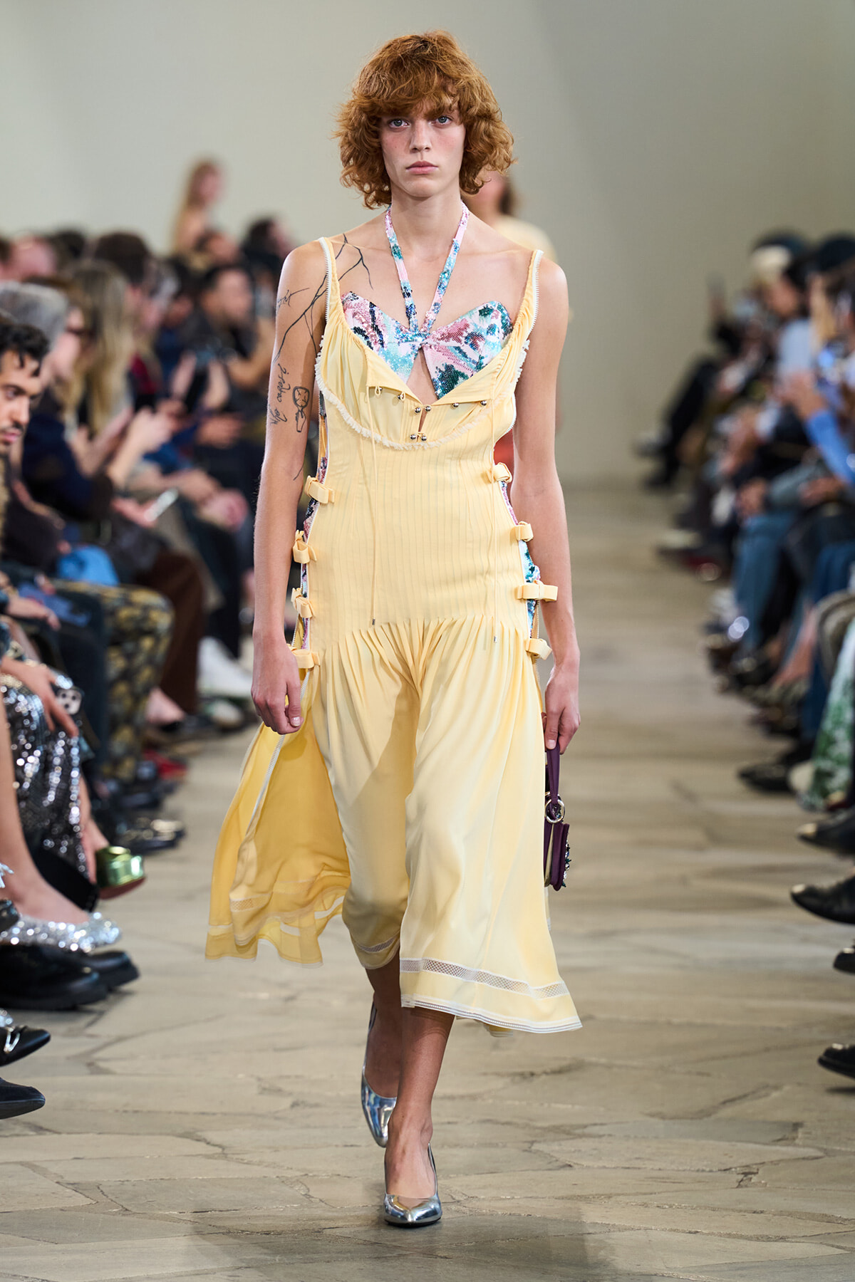 Model walking a runway in a pale yellow dress with a floral bustier top and side buckle details, audience blurred in the background.