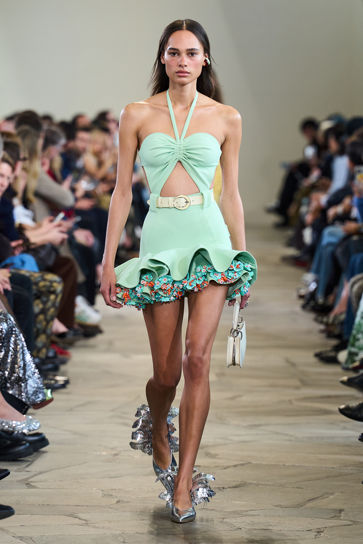 Model wearing a mint green halter dress with ruched bodice and cutout, high-waisted skirt with a floral-embellished ruffle, walking a runway.