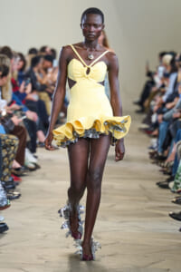Model walking a fashion show runway in a pale yellow, sleeveless dress with cutouts and a voluminous ruffled hem; metallic leaf-embellished heels visible on the floor.