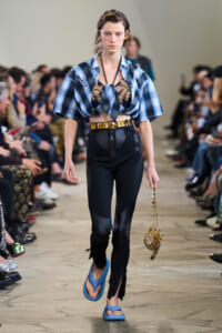 Model walks a runway in a blue plaid knotted crop top, black high-waisted pants, and blue sandals, holding a gold chain purse.