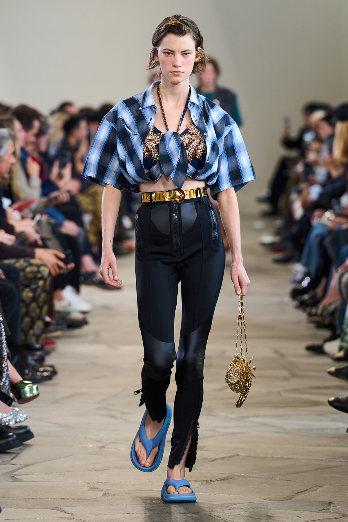 Model walks a runway in a blue plaid knotted crop top, black high-waisted pants, and blue sandals, holding a gold chain purse.