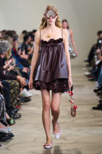 Runway model in a burgundy satin mini dress with a ruffled bodice and floral-embellished hem, wearing oversized goggles.