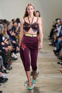 Model on a runway wearing a burgundy leather bralette, high-waisted maroon pants with red floral embroidery, a pale yellow belt, and turquoise fringe heels.