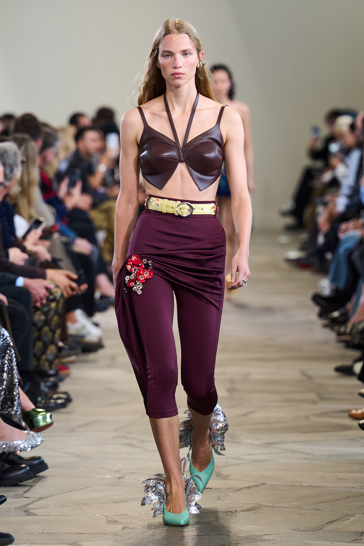 Model on a runway wearing a burgundy leather bralette, high-waisted maroon pants with red floral embroidery, a pale yellow belt, and turquoise fringe heels.