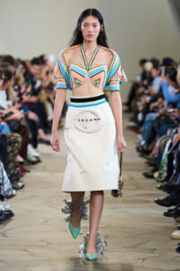 Runway model wearing a pastel geometric crop top and white midi skirt with a large circular belt buckle, mint heels with metallic fringe.