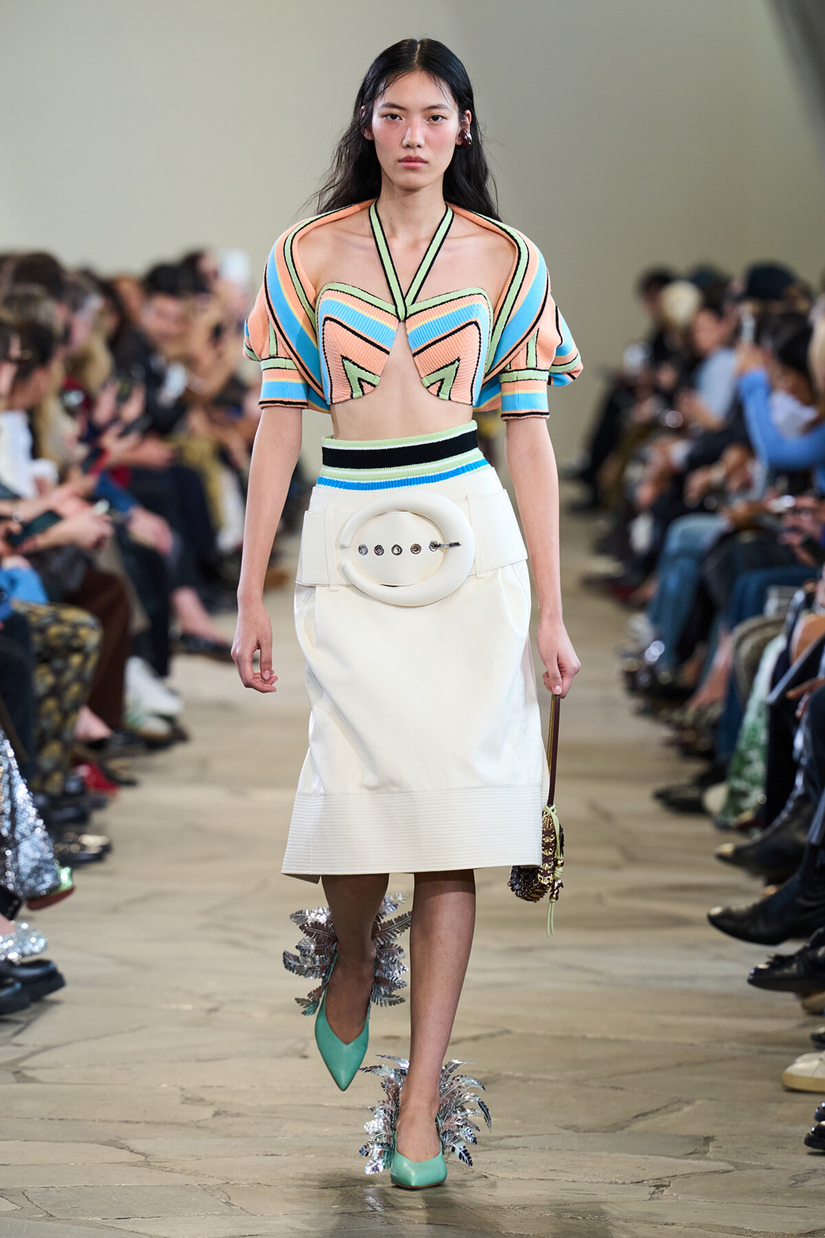 Runway model wearing a pastel geometric crop top and white midi skirt with a large circular belt buckle, mint heels with metallic fringe.