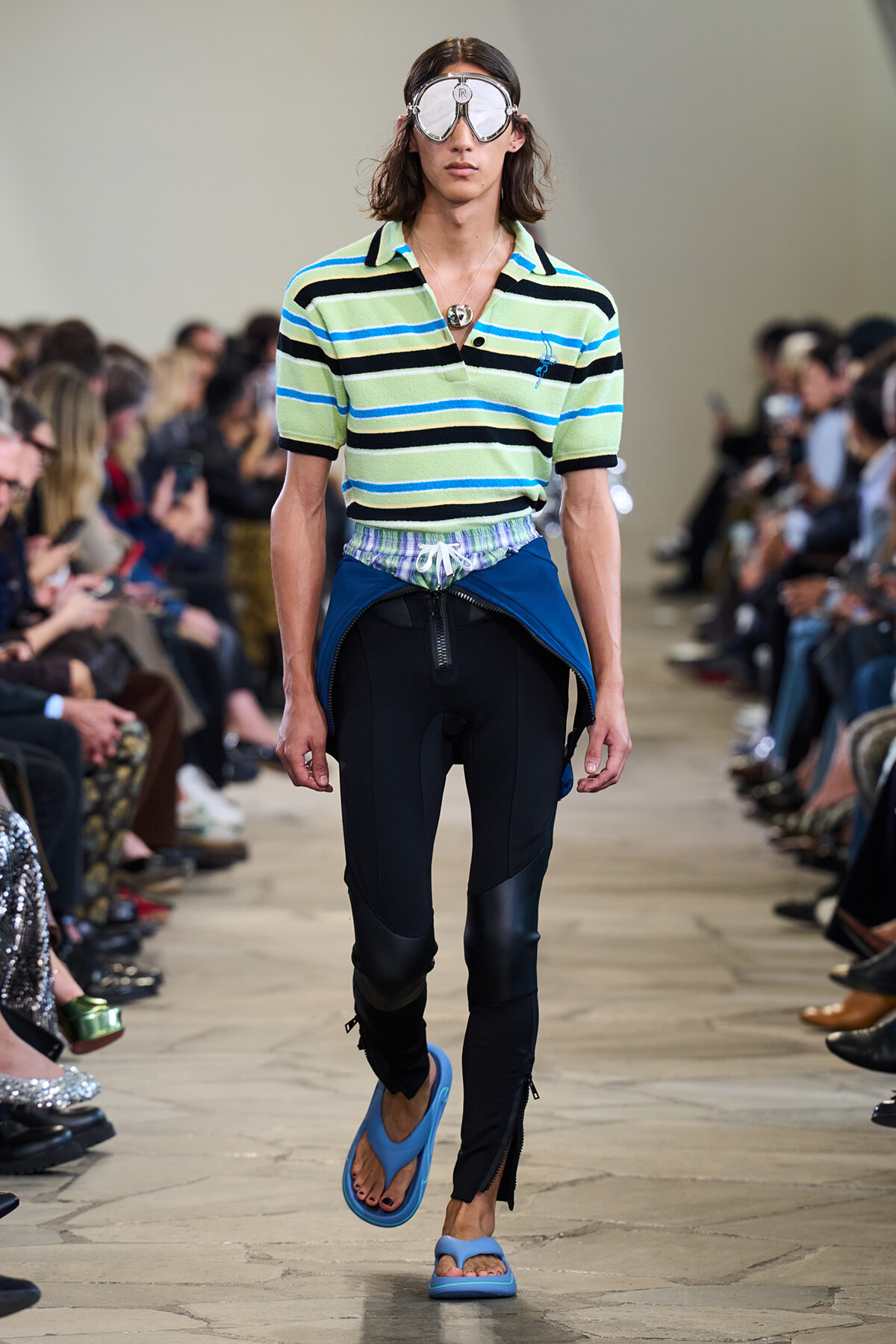 Male model walking a runway in a striped polo, goggles, and blue sandals; audience seated along the sides.