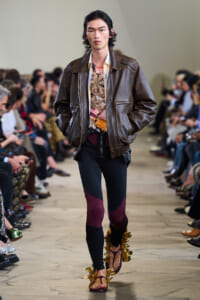 Male model walking a fashion show wearing a brown leather bomber jacket over a printed top and color-blocked pants, with gold decorative sandals and audience watching from the sides.