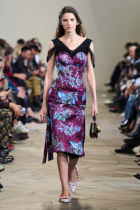 Model walking on a runway in a purple tropical-print dress with sheer black shoulder detailing, holding a small black handbag with gold accents.