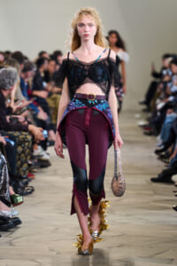 Runway model wearing a sheer black off-shoulder top over a blue floral bra, burgundy color-block pants with black panels, floral belt, gold spike ankle details, silver beaded bag, audience in background.