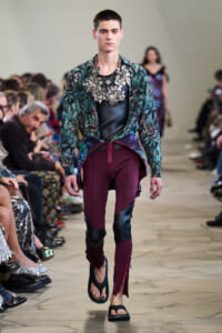 Male model on a runway wearing a tropical print shirt over a black top, a large metallic floral necklace, and burgundy panel pants with zippers adds bold flair to the look.