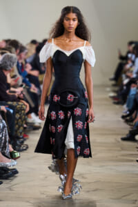 Model walking a runway in a black corseted dress with sheer off-shoulder sleeves and red floral embroidery, carrying a round black purse and silver leaf shoes.