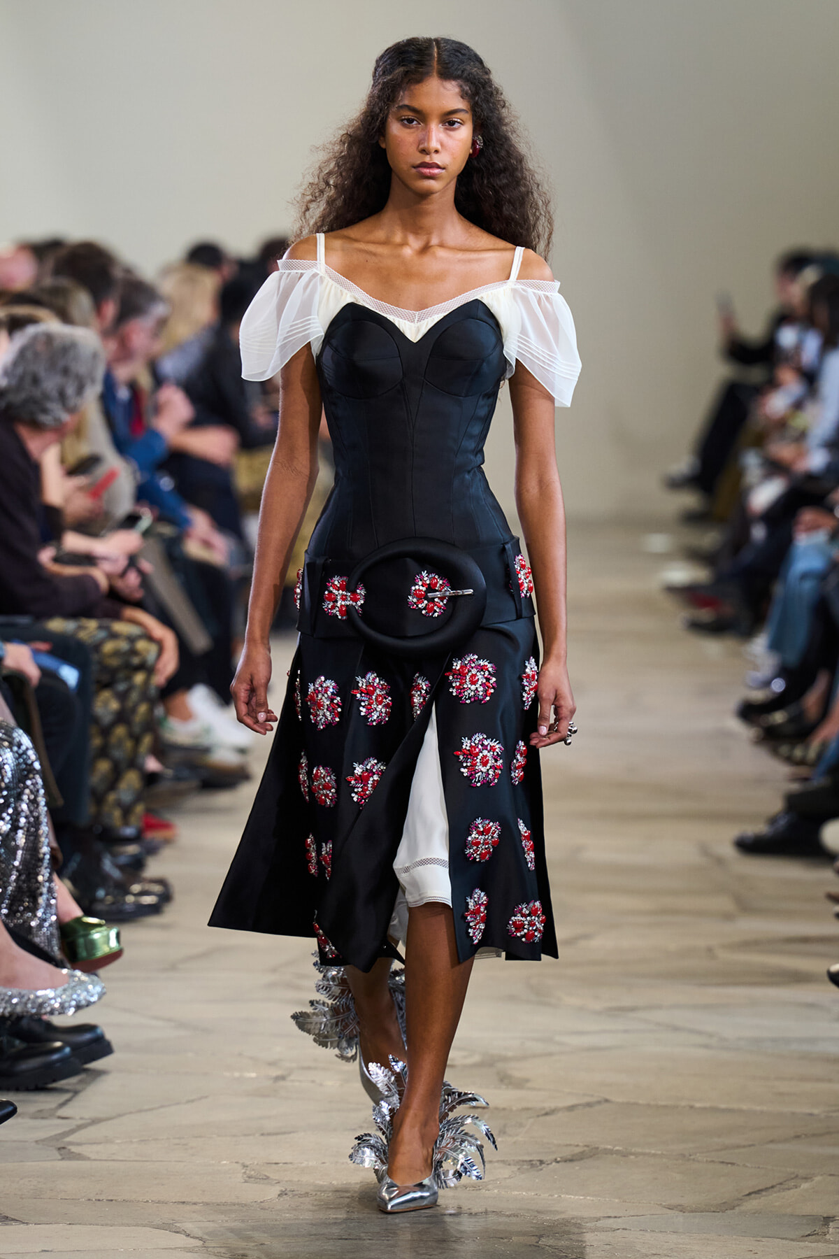 Model walking a runway in a black corseted dress with sheer off-shoulder sleeves and red floral embroidery, carrying a round black purse and silver leaf shoes.