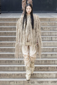 Model in a beige fringe ensemble walking down stone steps, long strands draping from shoulders to knees.