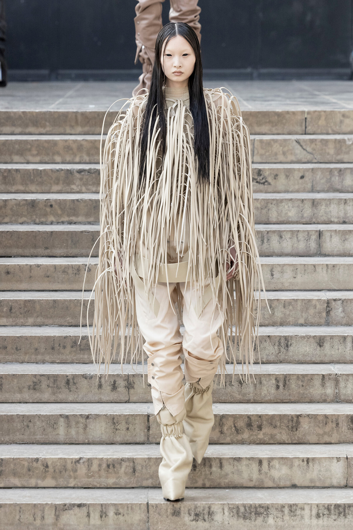 Model in a beige fringe ensemble walking down stone steps, long strands draping from shoulders to knees.