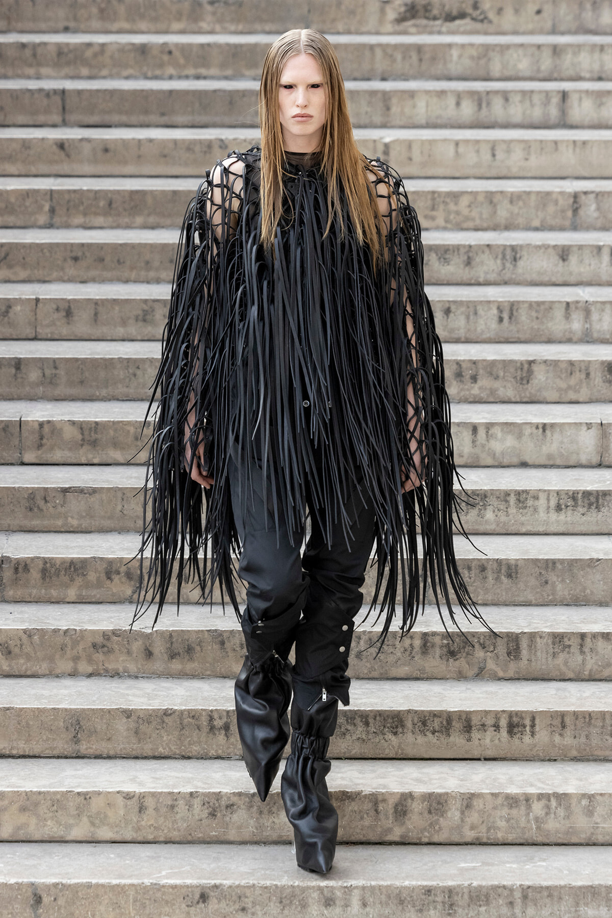 Model wearing a black fringe cape-like garment and tall black boots standing on concrete steps.