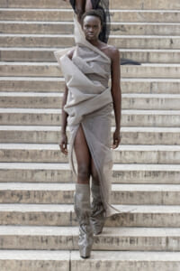Runway model in a sculptural gray draped dress with a high slit, descending stone stairs in matching gray boots.