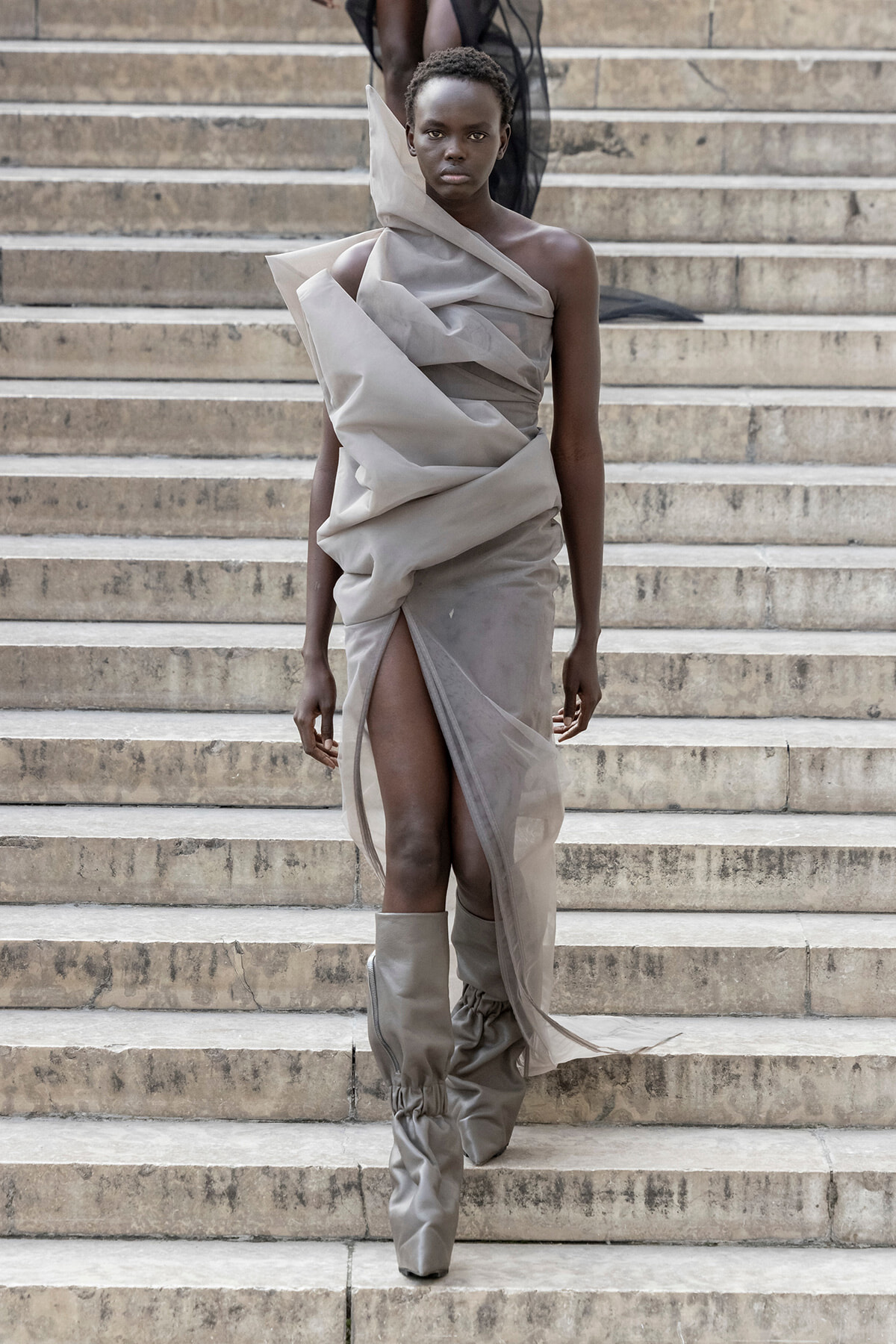 Runway model in a sculptural gray draped dress with a high slit, descending stone stairs in matching gray boots.