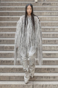 Person wearing a light gray fringe coat with long hanging ribbons standing on concrete steps in a fashion pose.