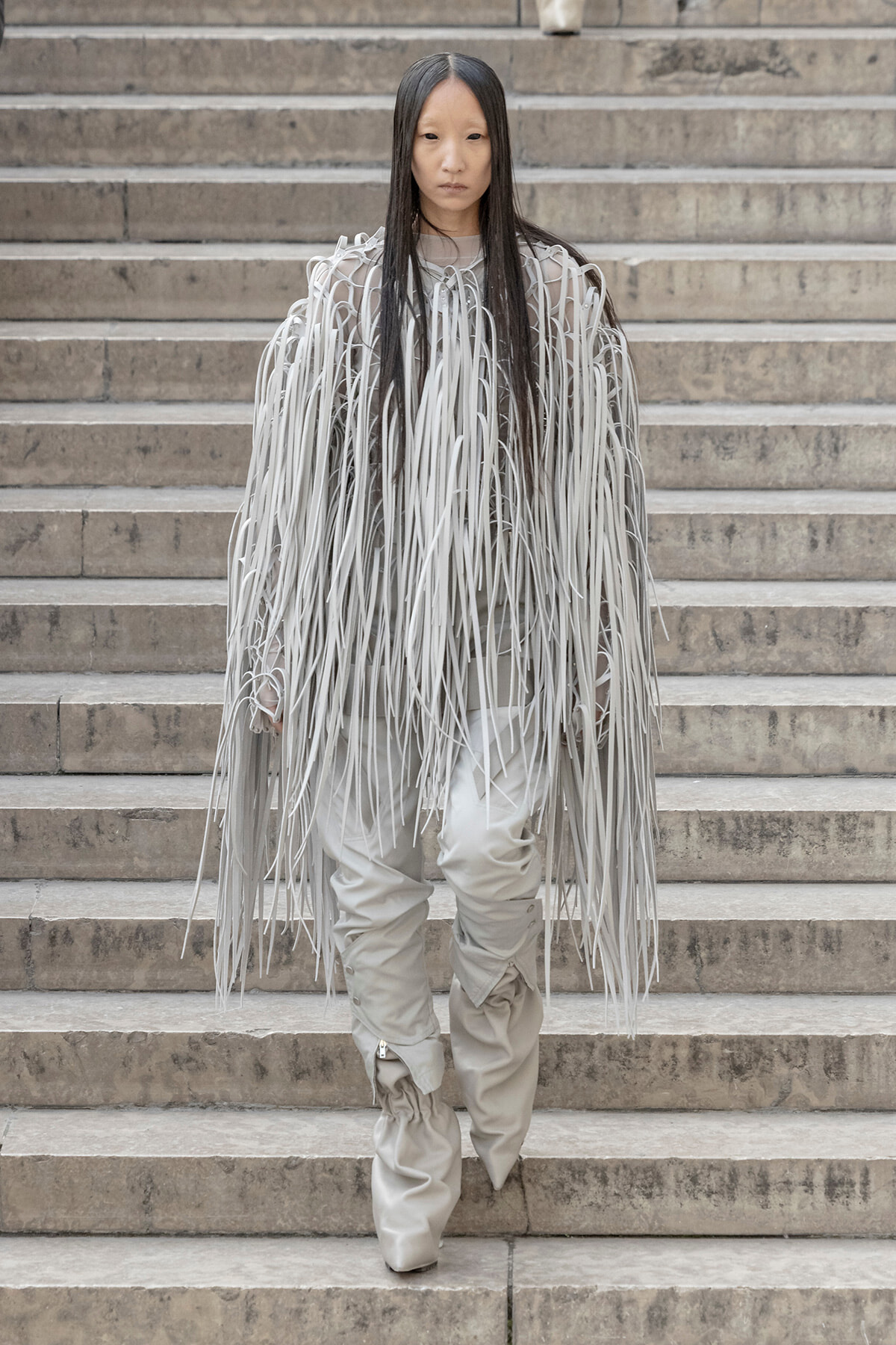 Person wearing a light gray fringe coat with long hanging ribbons standing on concrete steps in a fashion pose.