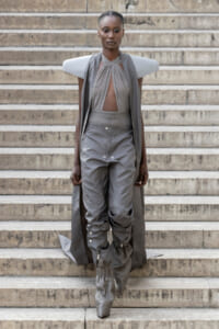 Model wearing a gray avant-garde outfit with sculptural shoulder panels and cape on stone stairs, mid-step downwards.