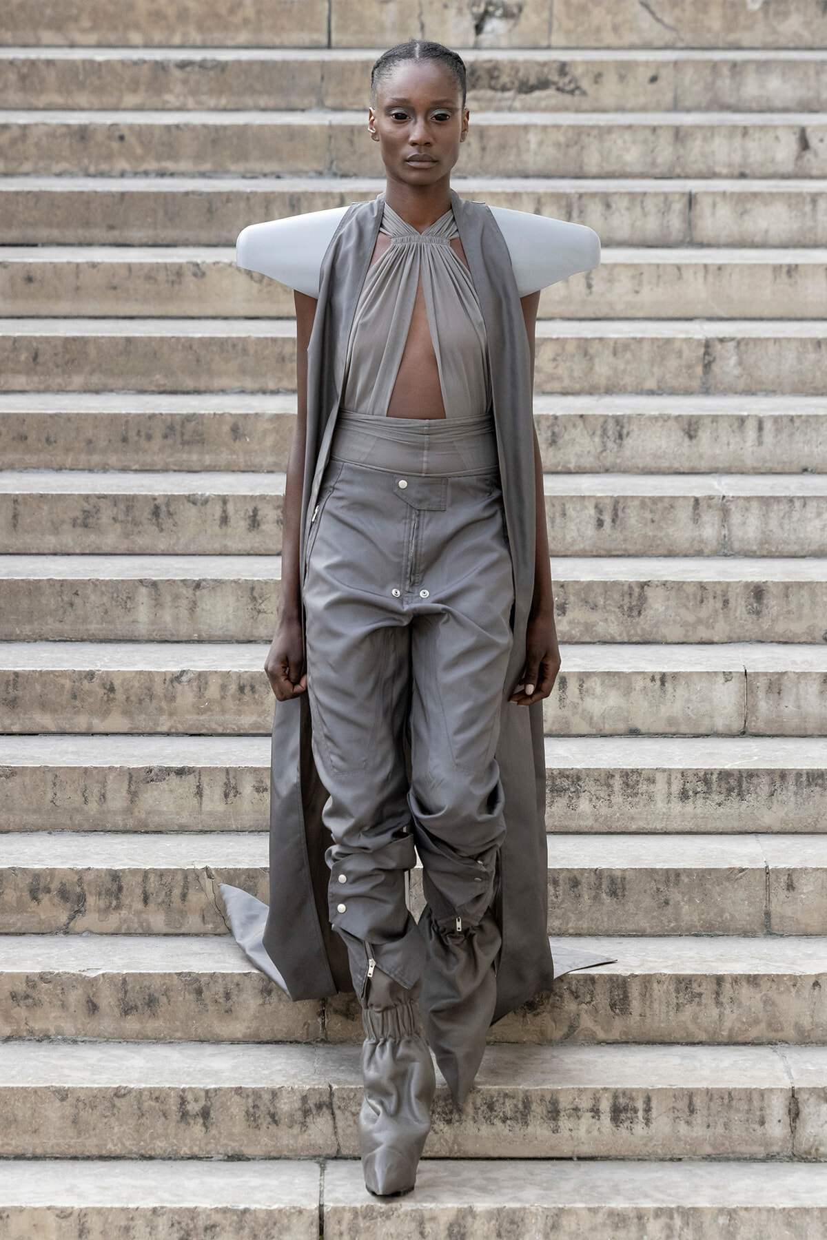 Model wearing a gray avant-garde outfit with sculptural shoulder panels and cape on stone stairs, mid-step downwards.