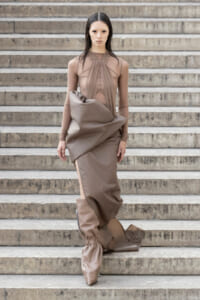 Model in a taupe sheer top and sculptural draped skirt walking down stone stairs with a monochrome, avant-garde look.