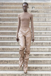 Model walking down stone stairs in a nude mesh long-sleeve top and tan pants with ruched knee-high boots on each leg.