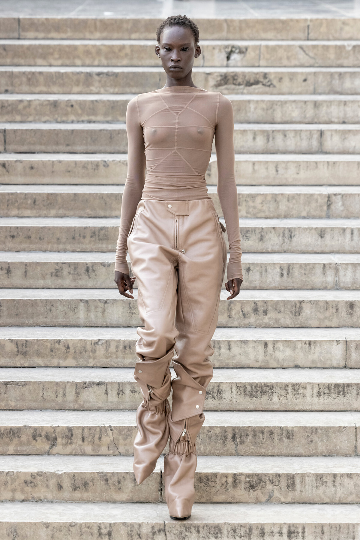 Model walking down stone stairs in a nude mesh long-sleeve top and tan pants with ruched knee-high boots on each leg.