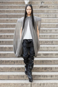 Model in a futuristic gray cape over a white top and black structured pants stands on stone steps, looking forward.