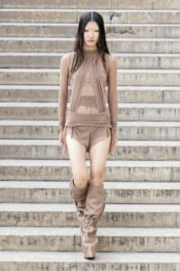 Young woman in a taupe sheer dress and slouchy thigh-high boots walking down stone steps, fashion-forward pose.