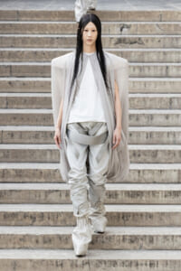 Model wearing an oversized light gray cape over a white top and sculptural pale pants as she walks down stone steps (fashion runway).