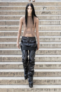 Model descending concrete steps in a sheer beige long-sleeve top and black strapped leather pants.