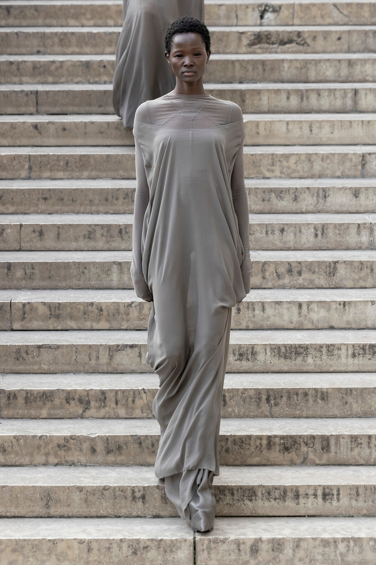 Woman models a loose, draped gray gown as she descends stone stairs.