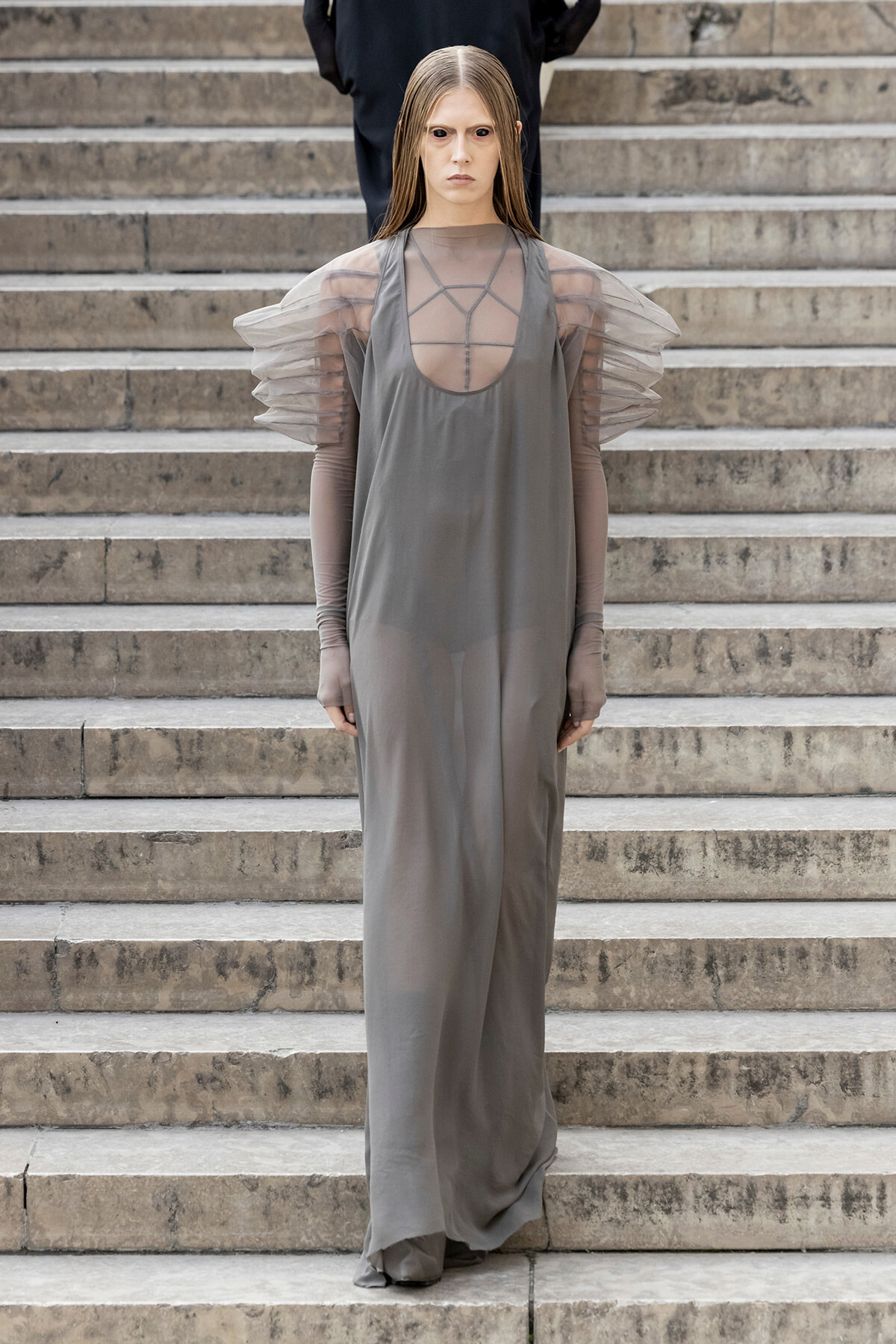Model descending stone stairs in a loose gray gown with sheer puff sleeves and geometric neckline.