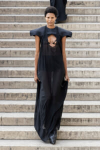 Model wearing a long black dress with dramatic shoulder pads descending a stone staircase.