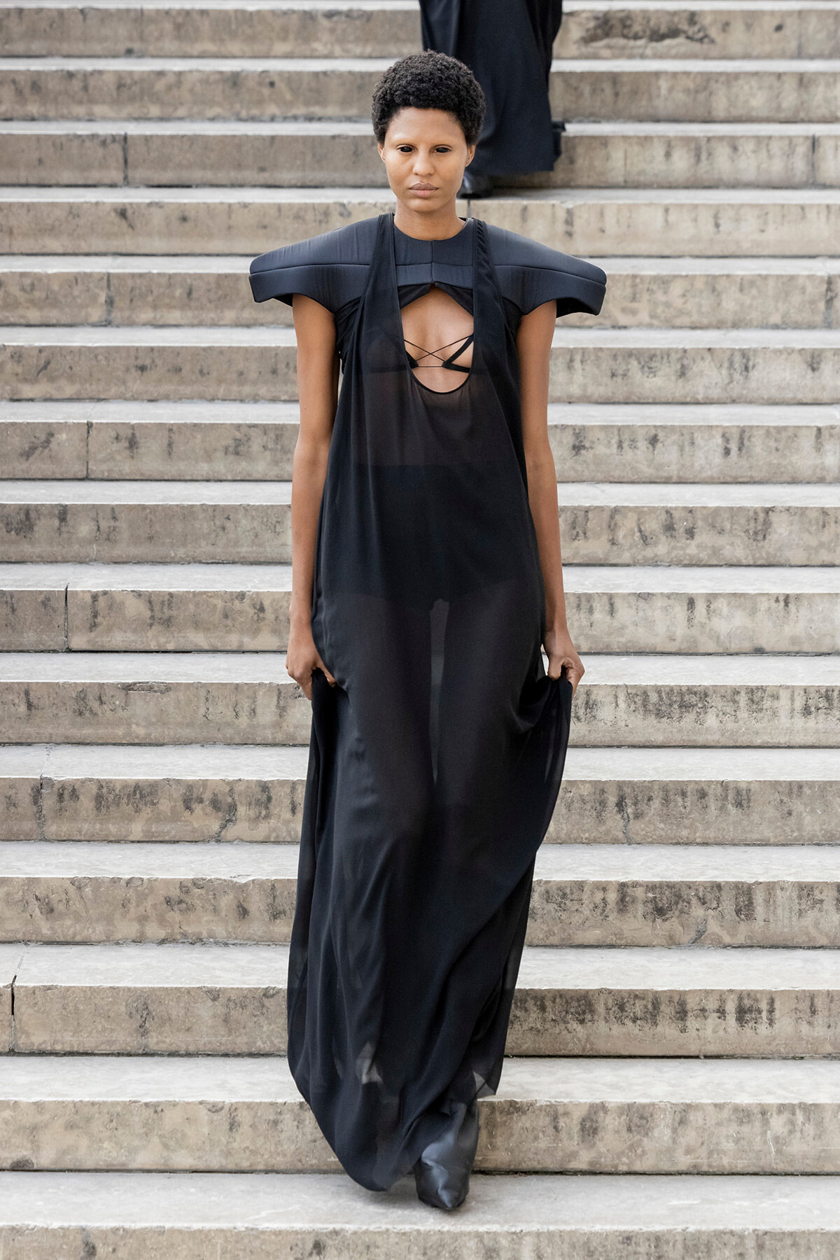 Model wearing a long black dress with dramatic shoulder pads descending a stone staircase.