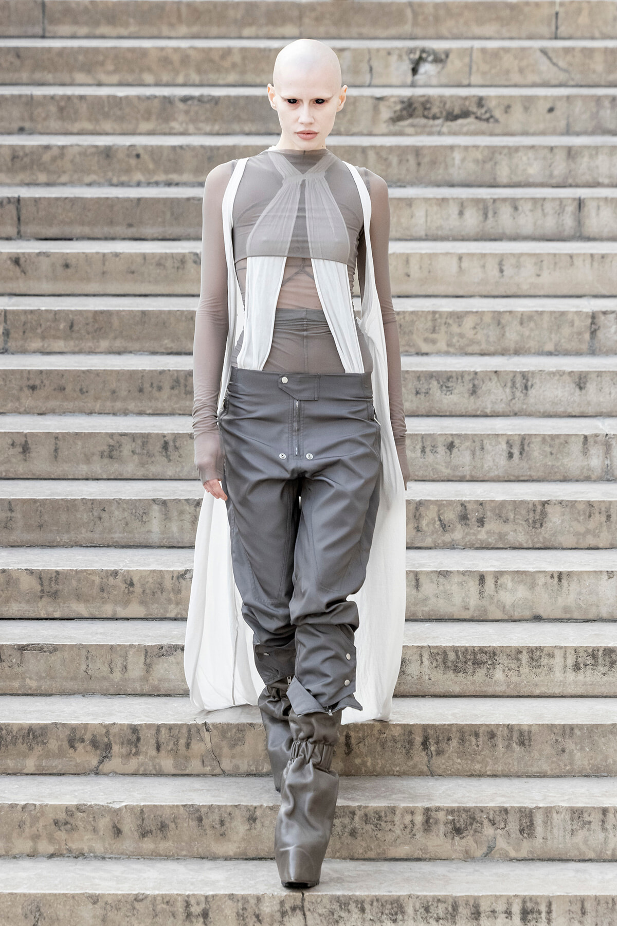 Fashion model walking down stone steps in a futuristic gray outfit: sheer top layers, white draped panels, and tall gray boots with gathered cuffs.