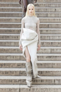 Model descending stone stairs in an ivory, draped dress with sheer sleeves and tall, light-beige boots.