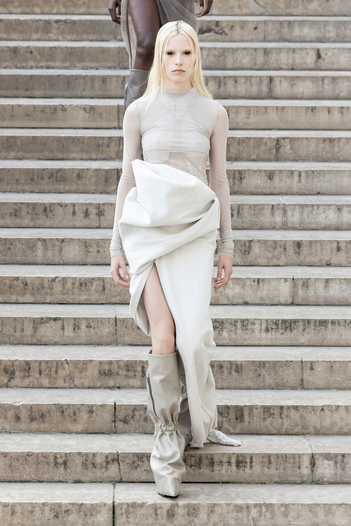 Model descending stone stairs in an ivory, draped dress with sheer sleeves and tall, light-beige boots.